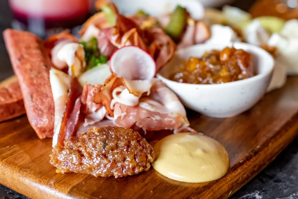 Close-up charcuterie board with sliced meats, radishes, pickles, cheese, and condiments.