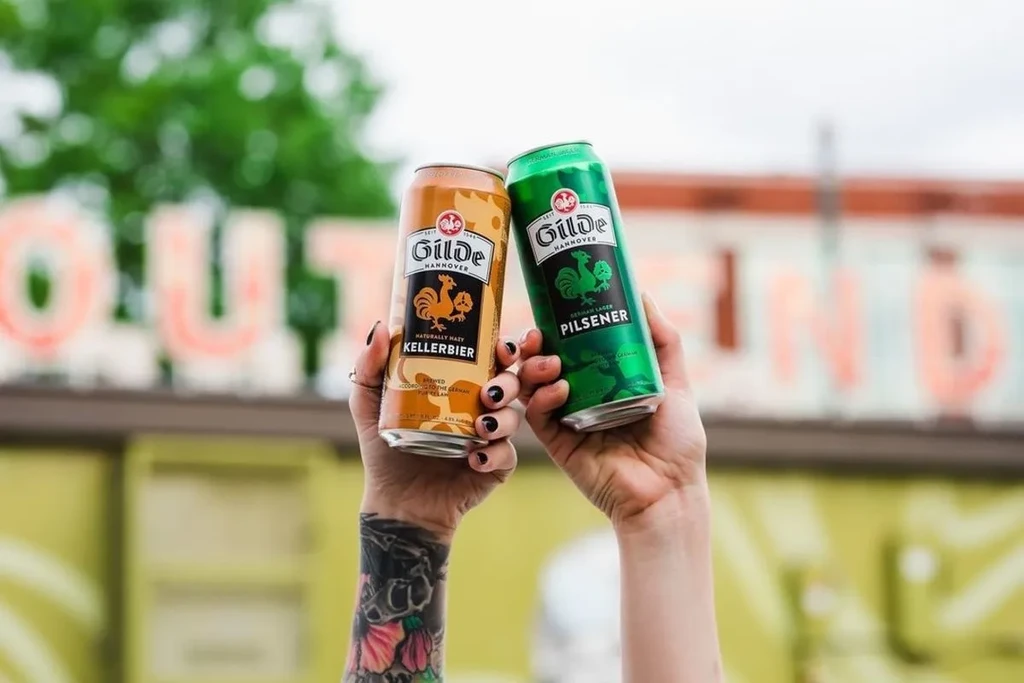 Two hands toast Gilde Hannover Kellerbier and Pilsener cans.