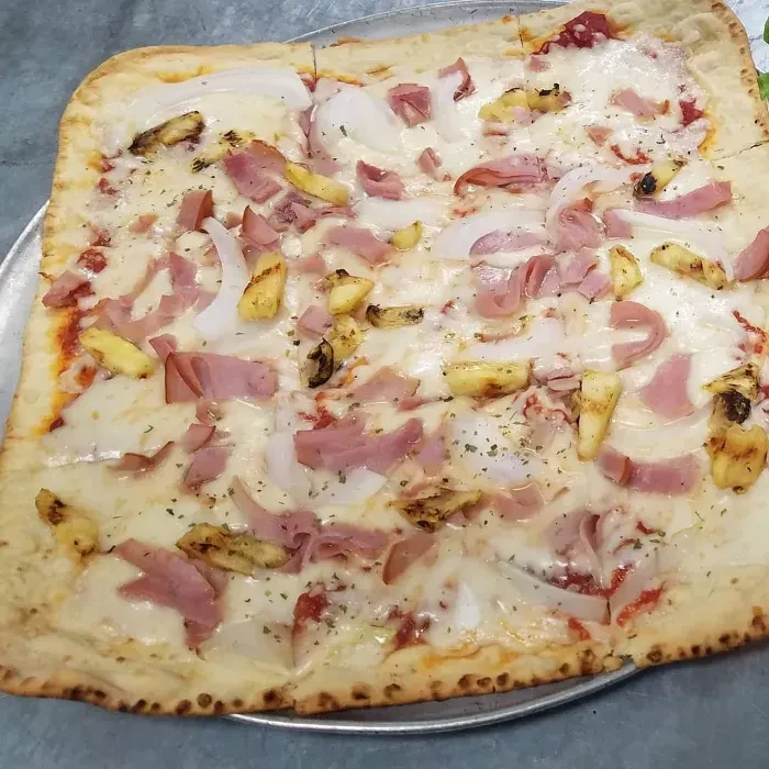 Rectangular pizza with cheese, ham, onion, pineapple, and herbs on a pan.