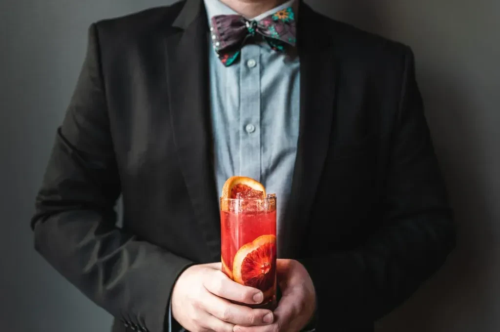 Person in suit and bow tie holds red cocktail with citrus slices.