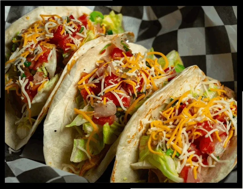Three soft-shell tacos with lettuce, tomato, cheese on checkered paper.