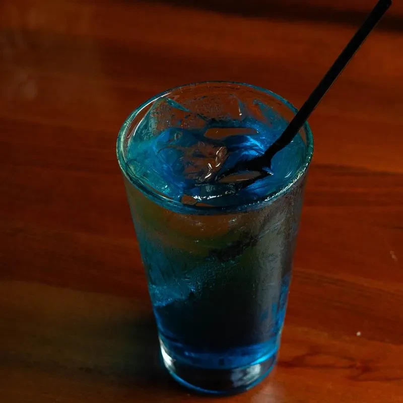 Tall glass of cold blue drink with ice, straw, on dark wood.