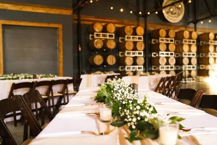 Warm event space with long tables, floral centerpieces, and wooden barrels.