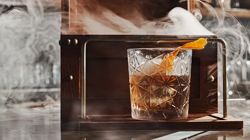Cocktail, ice, orange peel, surrounded by smoke from a wooden cloche.