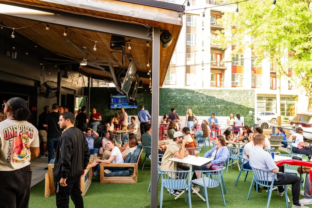 Bustling modern bar patio, half covered, half turf, filled with patrons.