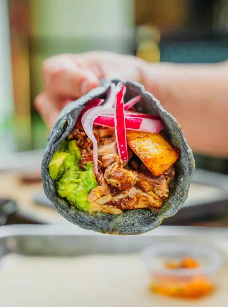 Hand holds a blue corn taco with meat, guacamole, and vibrant toppings.