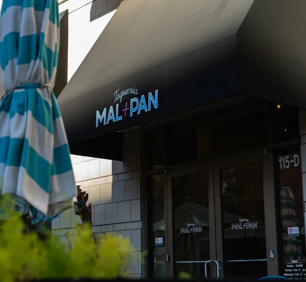 Taqueria MAL+PAN restaurant exterior with dark awning and striped umbrella.
