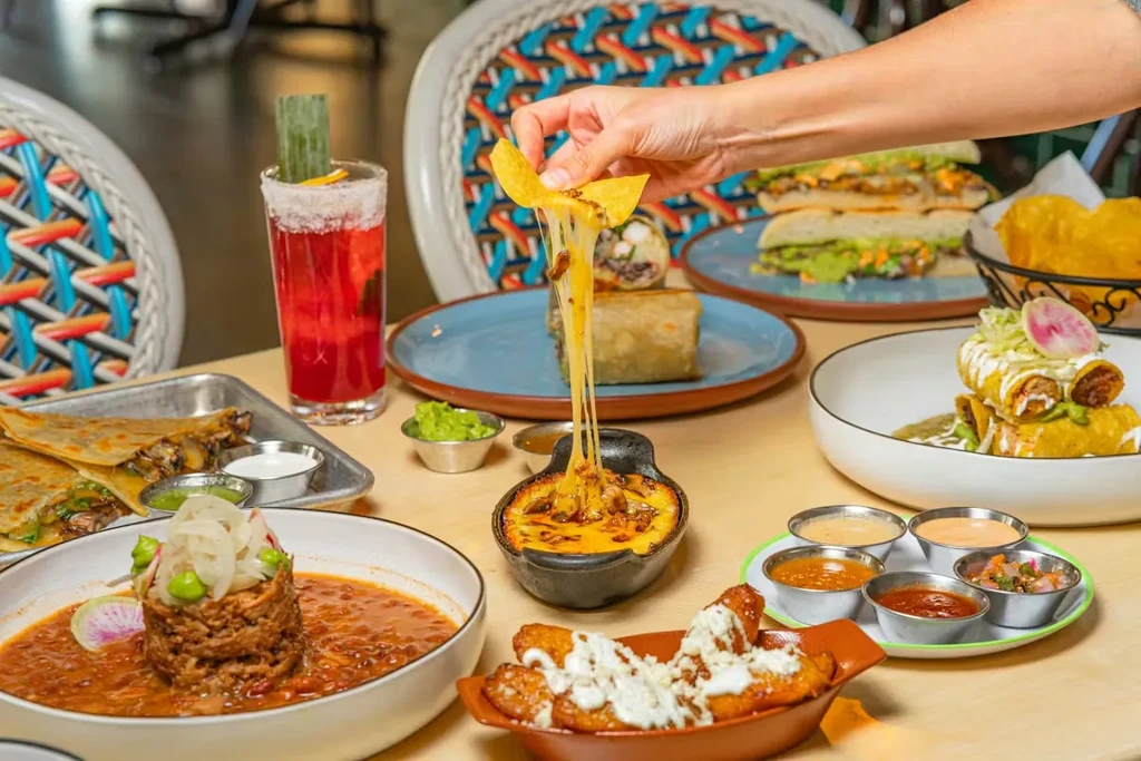 Vibrant table of Mexican food, hand lifting cheesy chip from queso fundido.