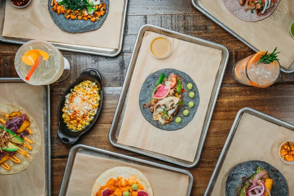 Assortment of colorful tacos, elote, and cocktails on a rustic table.