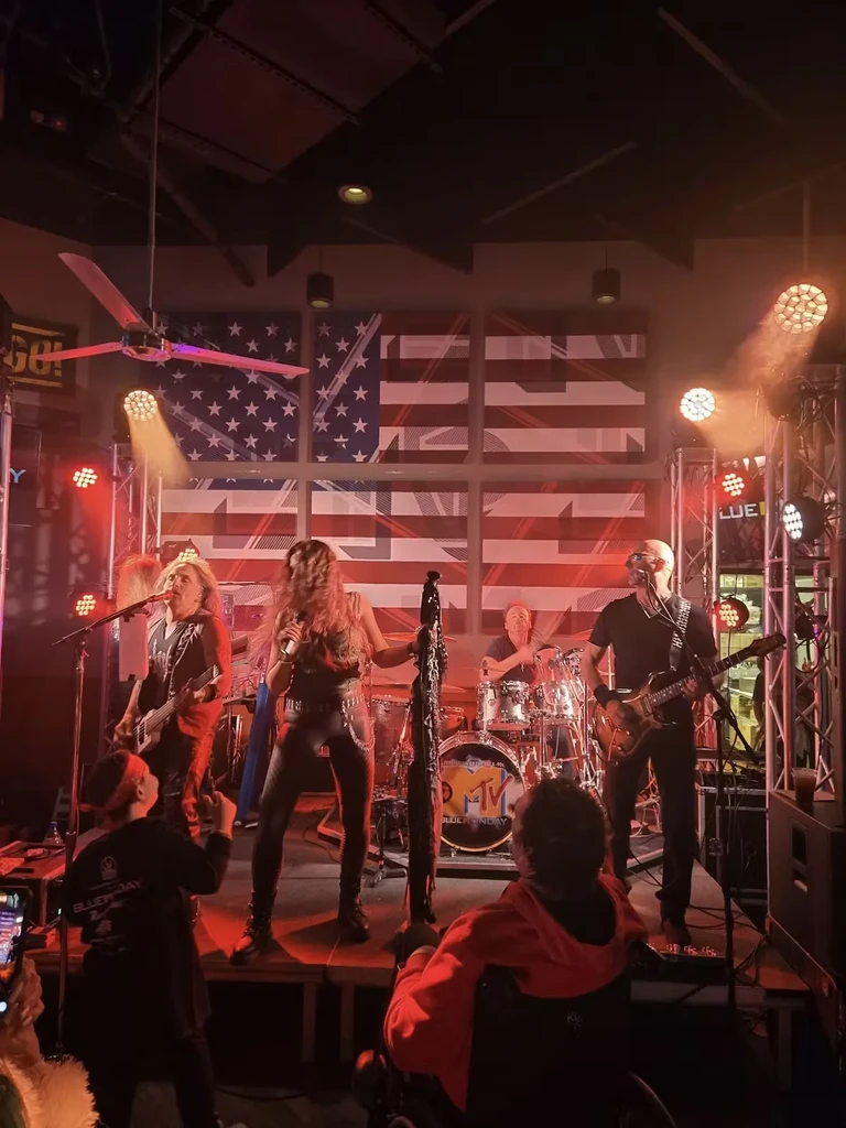 Live band performs on stage with flag backdrop, audience including wheelchair user.