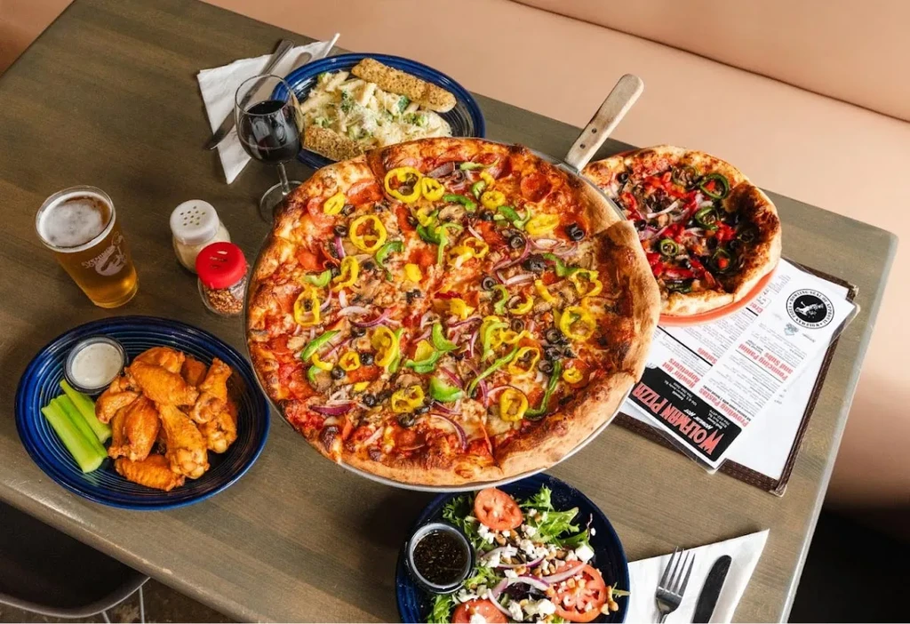 Restaurant table with two pizzas, wings, pasta, salad, beer, and wine.