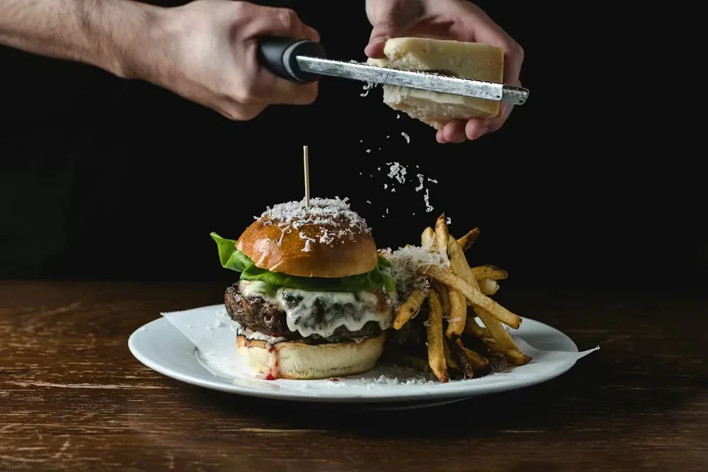 Hands grating cheese over a gourmet burger and french fries.