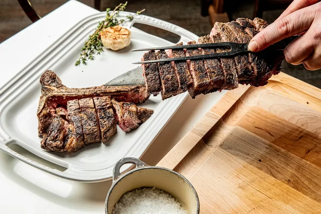 Hand serving pre-sliced steak, roasted garlic, and herbs from a platter.