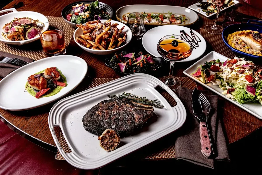 Dark table with steak, scallops, fries, salads, and cocktails.