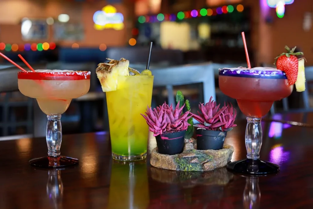 Three colorful cocktails on a bar with plants and blurred lights.