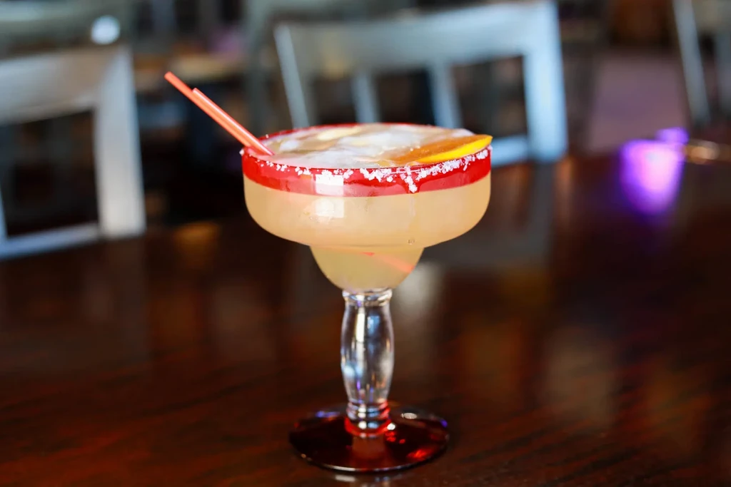 Stemmed cocktail with red salted rim, citrus wedge, blurred background.