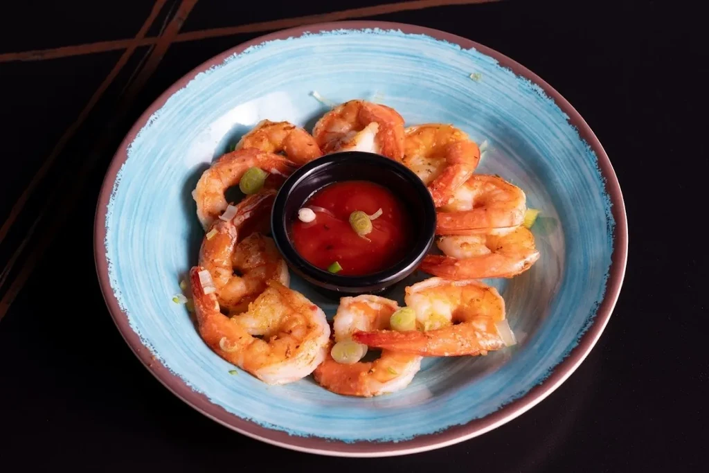 Cooked shrimp with green onions around red dipping sauce on blue plate.