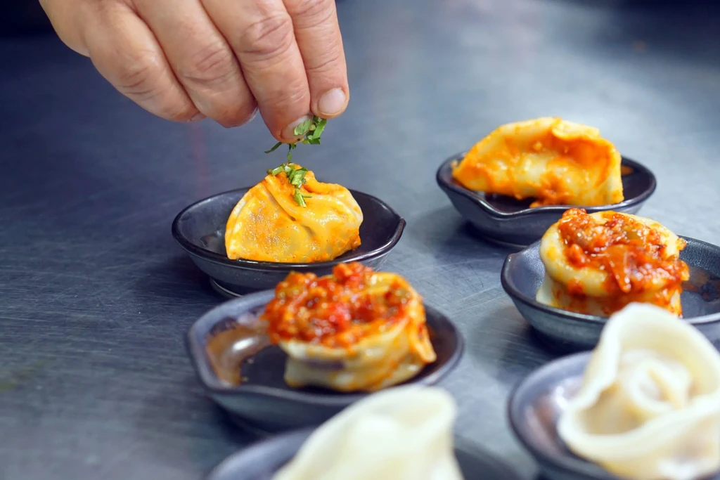 Hand sprinkles green herbs on red-sauced dumplings in dark dishes.