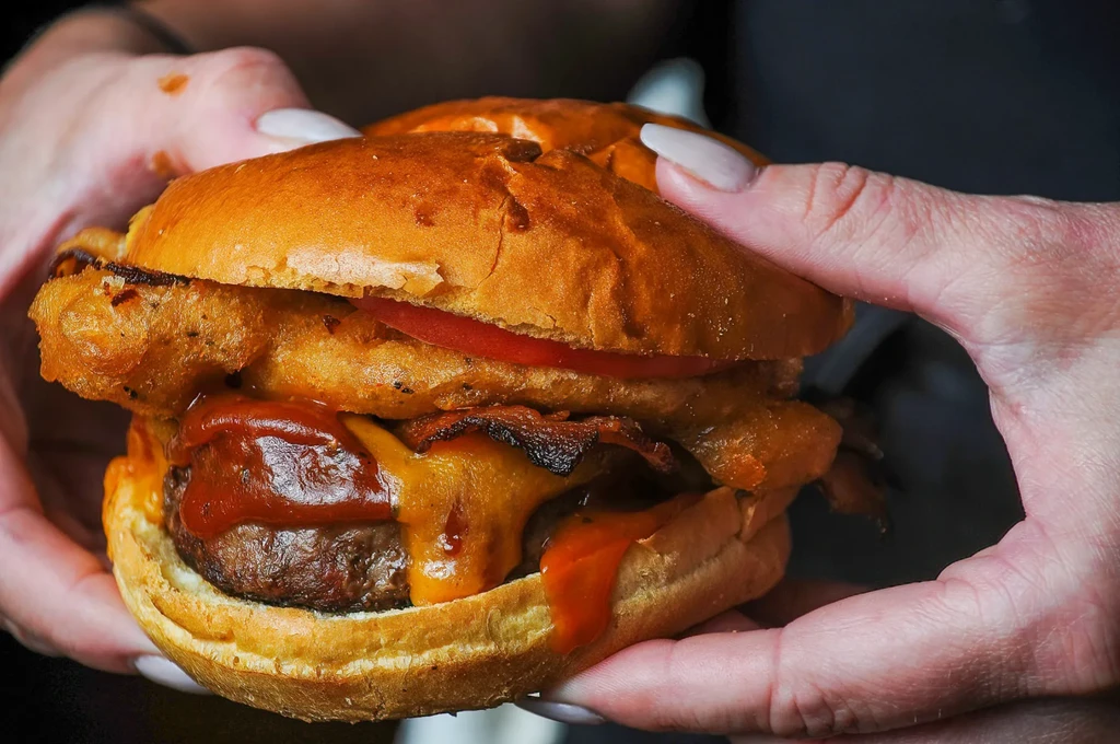 Two hands hold a large burger with patty, cheese, bacon, onion rings.