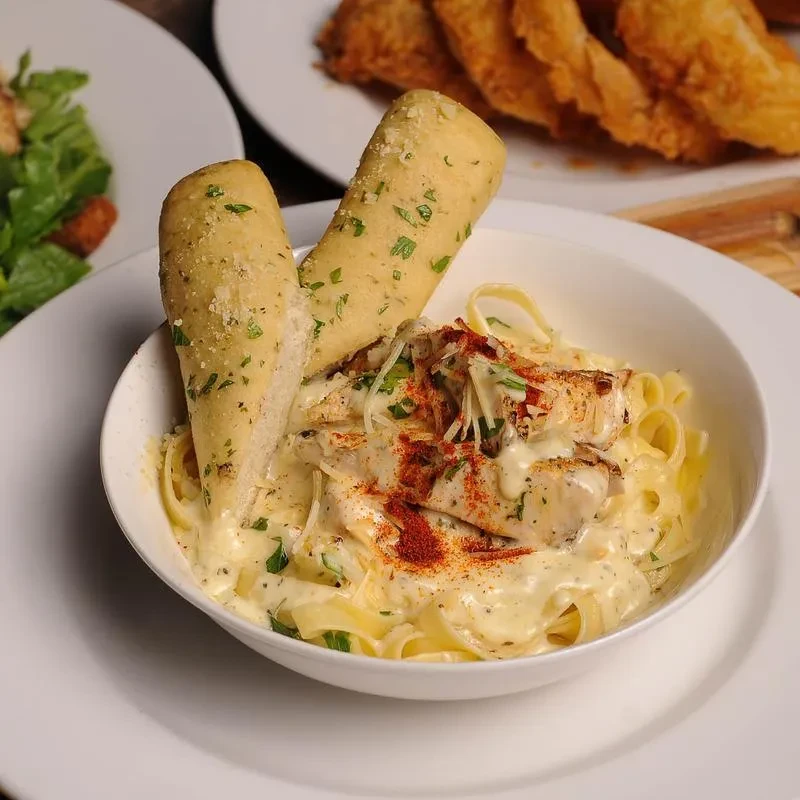 Bowl of creamy chicken fettuccine pasta with cheese, spice, and breadsticks.