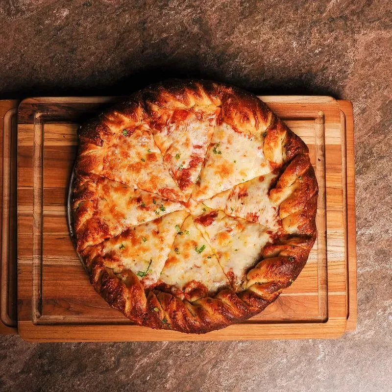 Freshly baked pizza with braided crust, melted cheese, herbs on a wood board.
