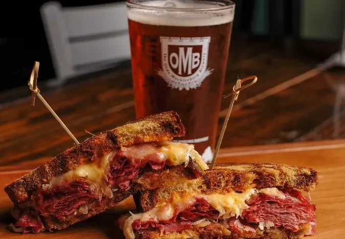 Sliced Reuben sandwich and OMB amber beer on a wooden surface.