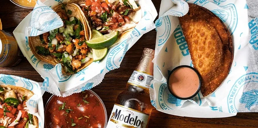 Overhead shot of Latin American tacos, empanada, salsa, and Modelo beer.