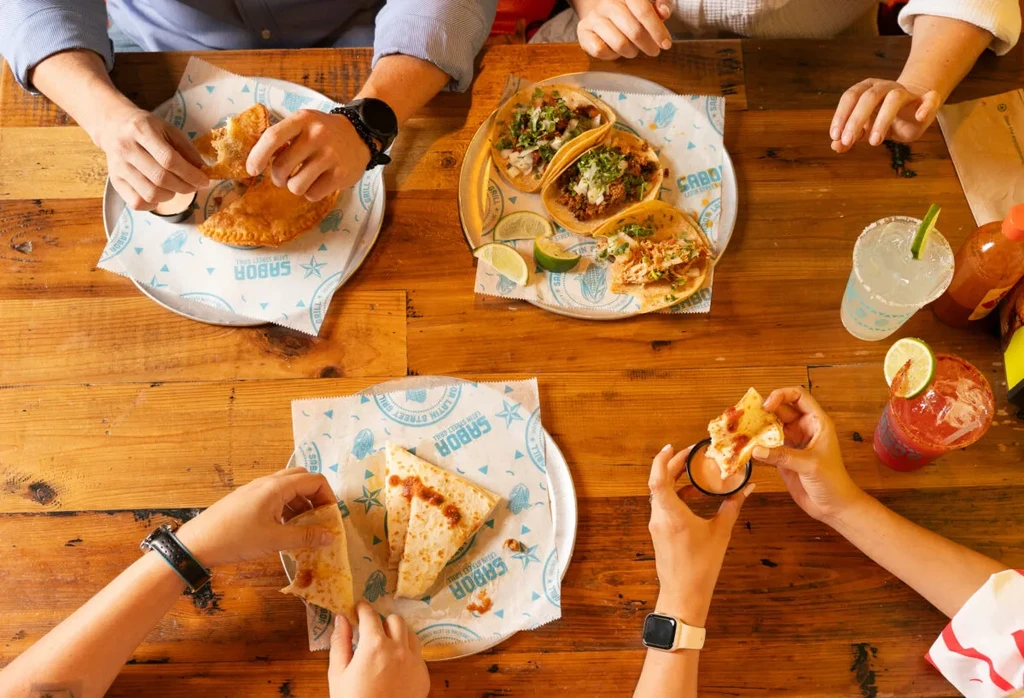 Overhead shot of hands sharing Latin street food and drinks.