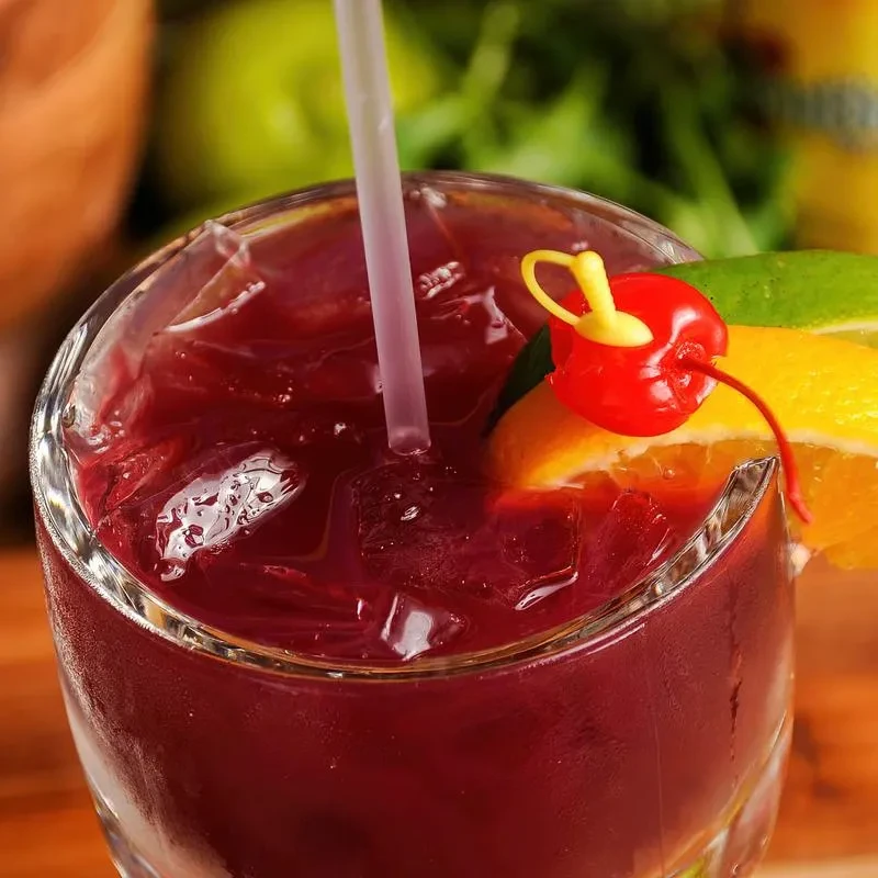 A reddish-purple cocktail with ice, straw, citrus slices, and maraschino cherry.