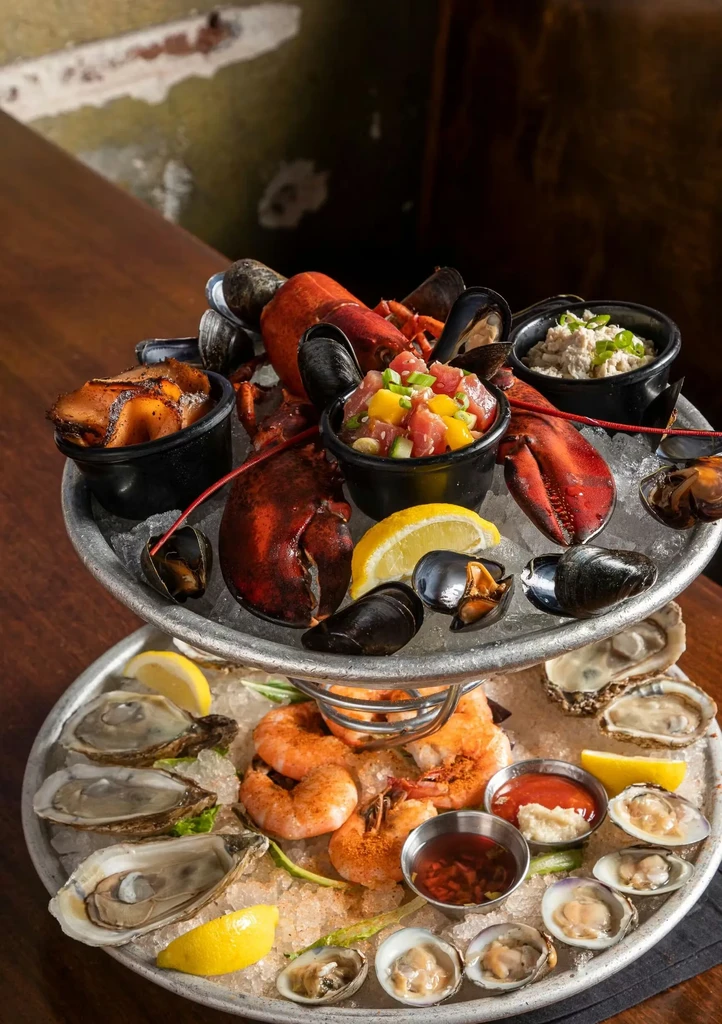 Two-tiered seafood tower on ice featuring lobster, oysters, shrimp, and poke.