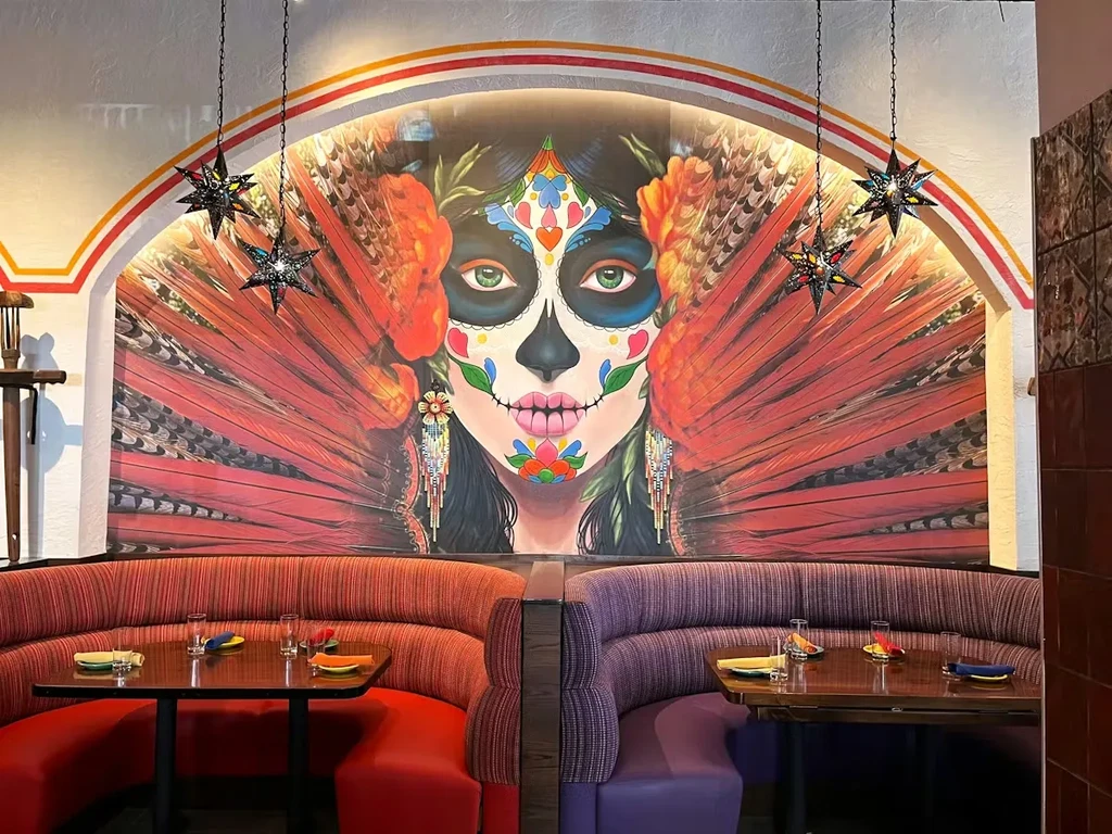 Colorful restaurant interior with large winged Calavera mural above booths.