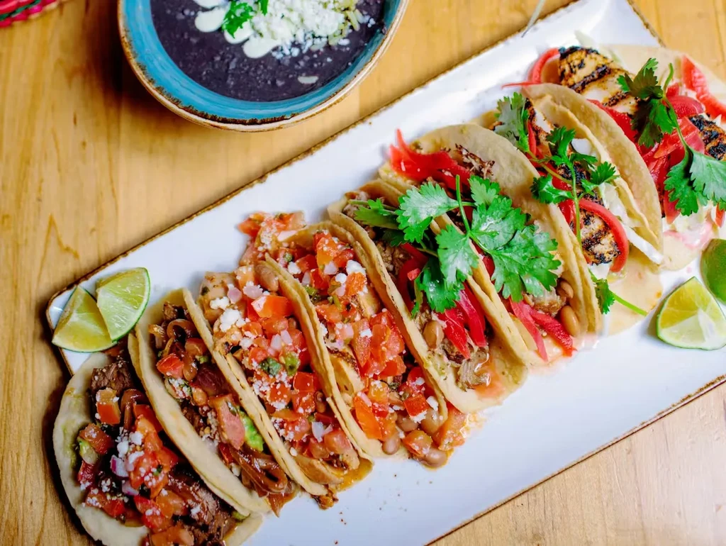 Eight colorful tacos with black bean soup on a rustic wooden table.
