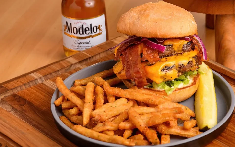 Double cheeseburger, fries, pickle, and Modelo Especial beer on wood.