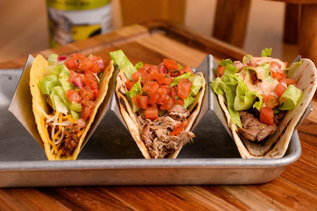Three distinct tacos in a metal holder on a wooden surface.