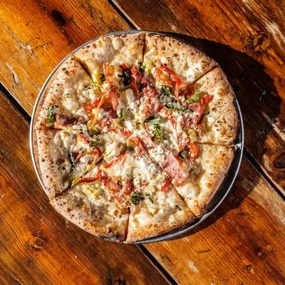 Rustic pizza with white sauce, cured meat, peppers, and herbs on wood.