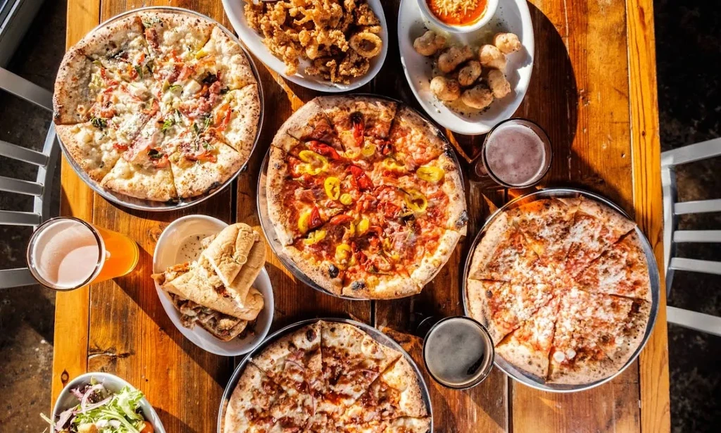 Rustic table overhead, laden with five pizzas, beers, and various sides.