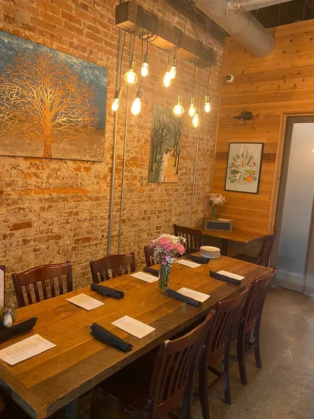 Rustic dining area with a long wooden table, flowers, and Edison bulbs.