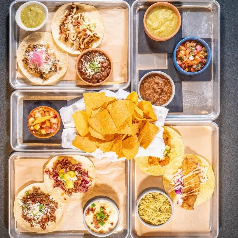 Overhead flat lay of Mexican tacos, chips, and dips on metal trays.