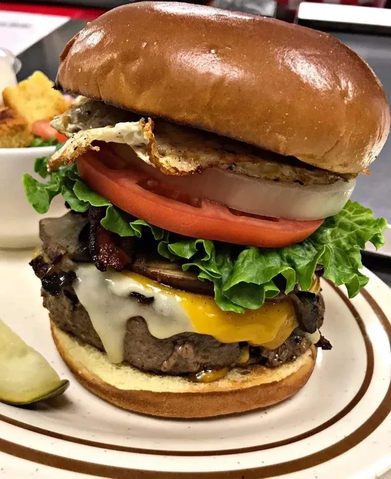 Stacked gourmet burger with egg, bacon, melted cheese, mushrooms, and vegetables.