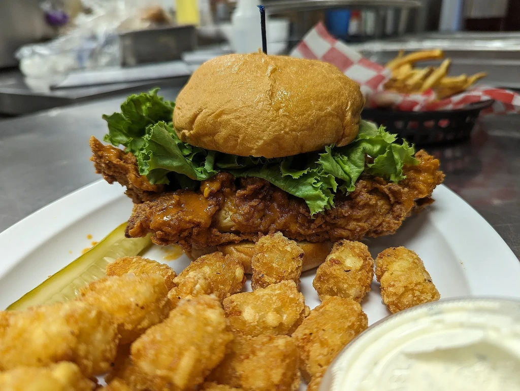 Close-up of a fried chicken sandwich, tater tots, pickle, and dipping sauce.