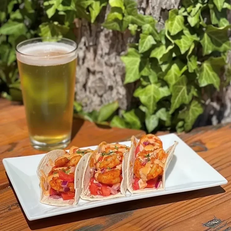 Three shrimp tacos and golden beer on a rustic table outdoors.