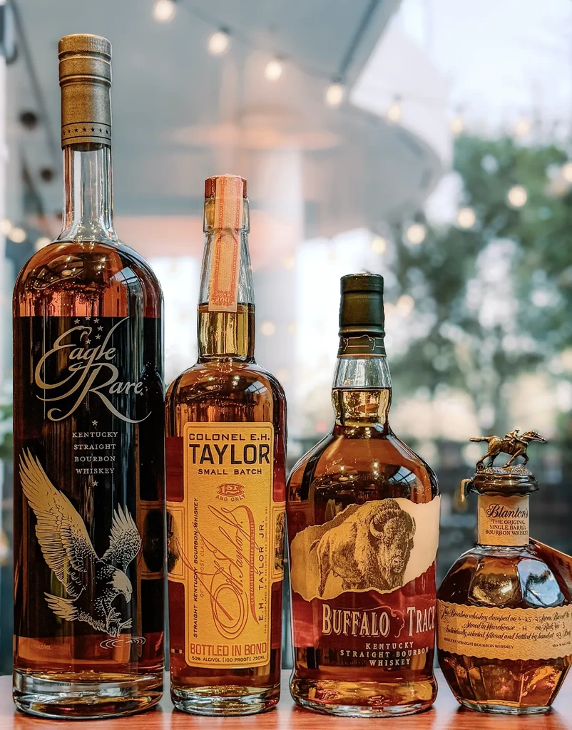 Four bourbon whiskey bottles, including Blanton's, on reflective surface.