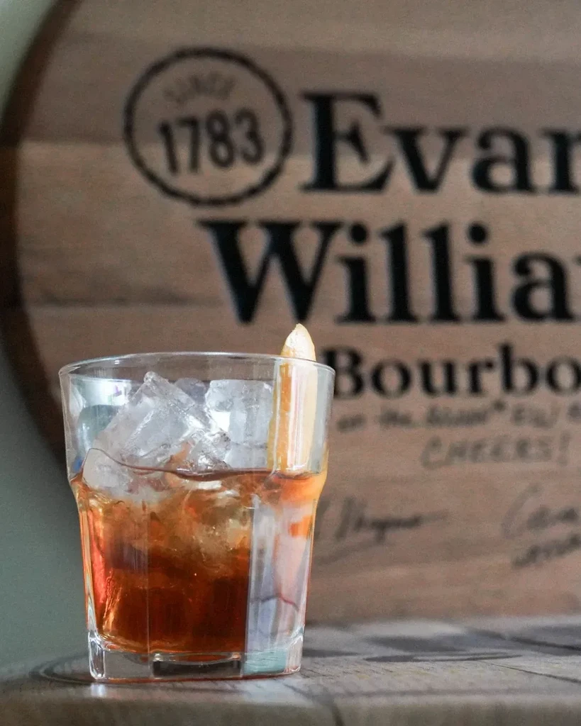 Rocks glass cocktail with orange peel, blurred Evan Williams bourbon barrel.