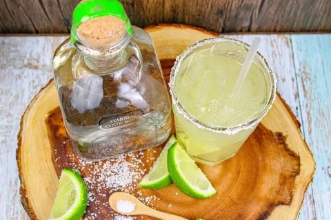 Tequila bottle, margarita, limes, and salt on a rustic wooden board.