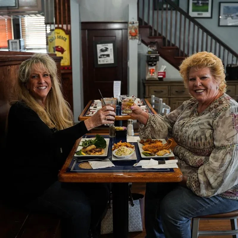 Two smiling women toast with drinks and food at a casual restaurant.