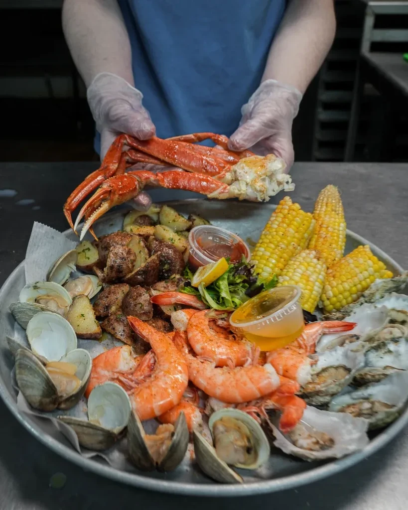 Gloved hands hold crab leg over cooked seafood and sides platter.