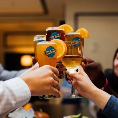 Hands clink Blue Moon beers garnished with oranges and other drinks.