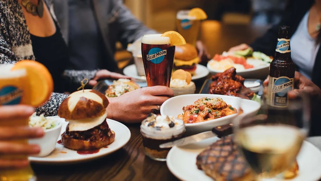 Bustling restaurant table with food, Blue Moon beer, and diners' hands.