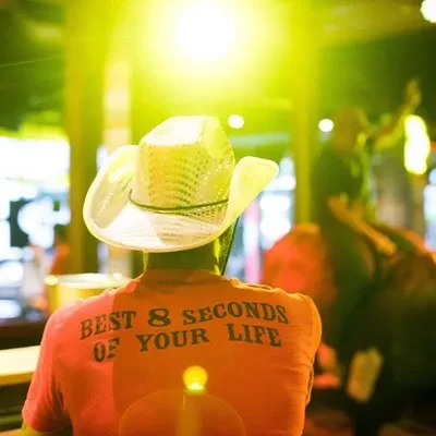 Person in cowboy hat, orange 'BEST 8 SECONDS' t-shirt, lively setting.