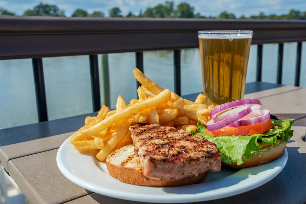 Grilled fish sandwich, fries, and beer served outdoors by water.